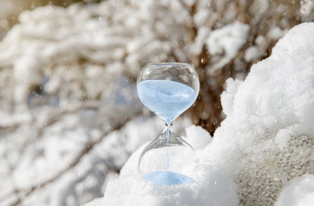 Conceptual image of switching to winter time or fall back, end of daylight saving time. Selective focus on hourglass with blue color sand falling in the winter in snowy nature.の写真素材