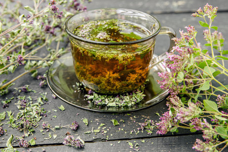 Homemade Oregano, Origanum vulgare herbal tea in a tea cup. Steaming hot drink with dry and fresh Oregano flower blossoms around, black color wood board minimal background.の写真素材