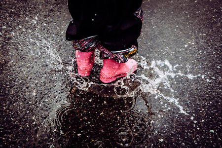 Detailed view of a young girl's feet having fun jumping in a water puddle on a wet street, wearing rain boots as water splashes.の写真素材