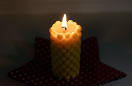A beeswax candle with a bubbly shape, cast in a handmade silicone mold, burns on a windowsill at home in dark.の写真素材