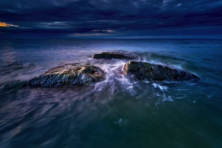 Wave-washed rocks stood out in the sea under a dramatic, dark evening sky. The long exposure creates...