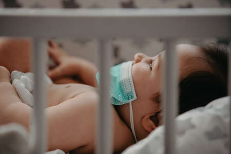 Sick baby with a mask on his face laying in his bed. Mother Checks The Temperature Of A Sick Baby.の写真素材