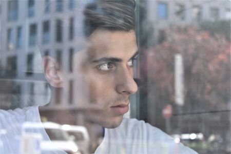 A thoughtful young man looks through a window in which the buildings of the city are reflectedの写真素材