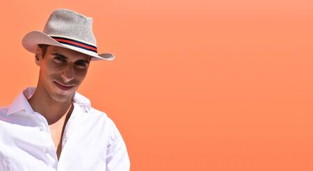 A smiling young man protects himself from the sun with a hat in front of a vivid orange backgroundの写真素材