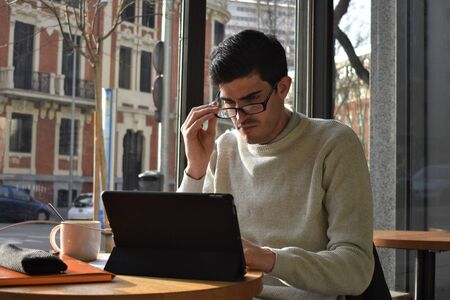 Side view of a young man putting on his glasses to work with a tablet while enjoying a cup of coffee in front of a windowの写真素材