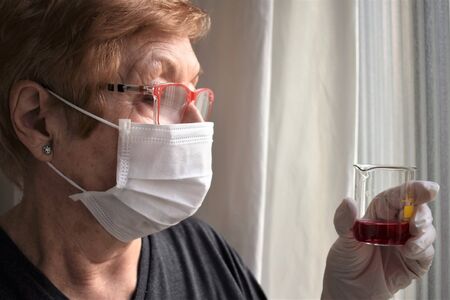 A lab scientist woman with gloves and protective mask observes a beaker with a red solution insideの写真素材