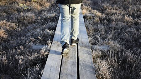 Detail of legs of a hiker wearing boots walking on a wooden walkway over frosty grass.の写真素材
