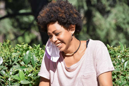Closeup of a young african origin woman, laughing with a face mask hanging in the ear, outdoors in a...