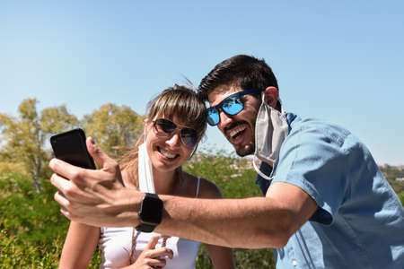A young couple, with removed face masks hanging from ear, using a smartphone for make selfie and...