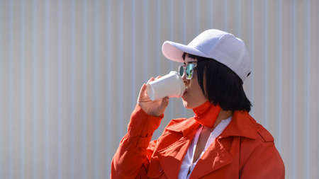 Portrait of a young Asian woman drinking coffee or tea while walking down the street with her face...