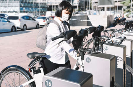 Young oriental woman with face mask takes a bike via renting services in a city center. Safe and...