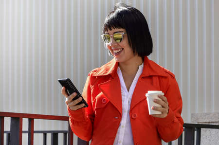 A young woman, with mirror sunglasses and a takeaway drink in her hand, smiles as she browses in her...