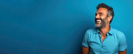 Handsome Indian man with blue shirt laughing on blue background.の素材