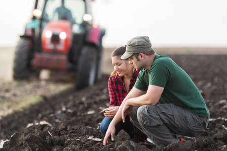 Young farmers examing dirt while tractor is plowing fieldの写真素材