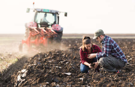 Young farmers examing dirt while tractor is plowing fieldの写真素材