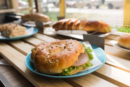 Fresh bun sandwich with ham, cheese, bacon, tomatoes, lettuce, cucumbers on light wooden table at window shopsの写真素材