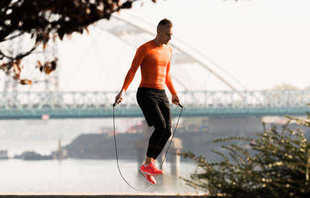 Handsome young man wearing sportswear and  skipping rope at quay during autumn