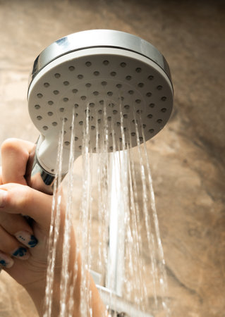 A woman's hand holds a shower head while taking a shower. Close-up of the hand and the stream of waterの写真素材