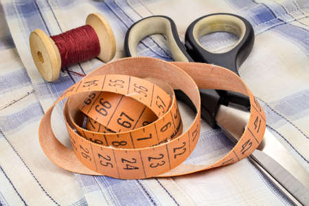 The tailor's tape measure, a wooden spool of thread and dressmakers scissors on fabric background
