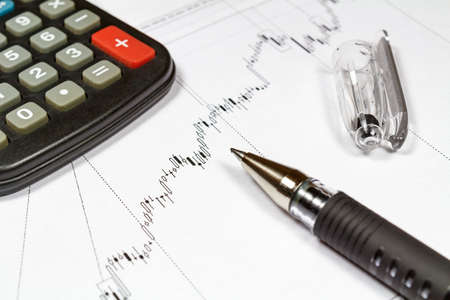 Fragment of electronic calculator and ball pen on the background of currency growth schedule