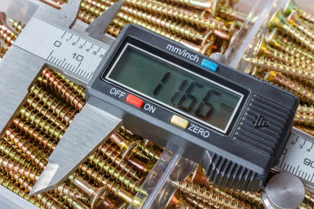 Digital electronic caliper on a background of opened storage box with yellow long screws closeup