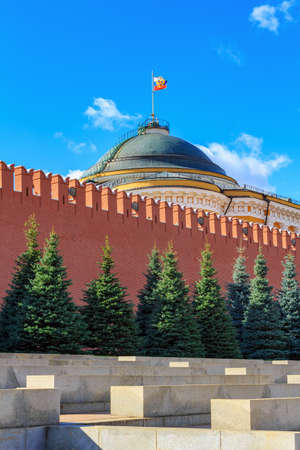 Moscow, Russia - September 30, 2018: View of Kremlin wall and green spruces against dome of Senatskiy Dvorets with Russian flag in sunny day on a background of blue sky with white cloudsのeditorial素材