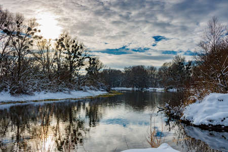River landscape in sunny winter day. Snow covered riversides of small river with trees and bushes in sunlight against blue sky with cloudsの写真素材