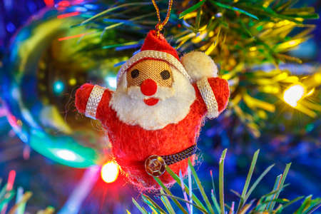 Handmade Santa Claus cloth figure on the branch of christmas fir against luminous colored garland...