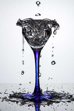 Full cocktail glass on blue thin and high leg with frozen water streams and flying water drops on dark glossy surface in white backlight. Stop motion photographyの写真素材