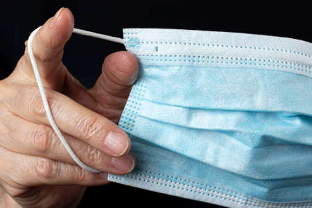 Blue disposable medical mask in wrinkled hand of elderly woman close-up on black background