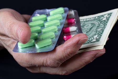 Two blister packs with colored pills and paper money in wrinkled hand of elderly woman on black background close-upの写真素材
