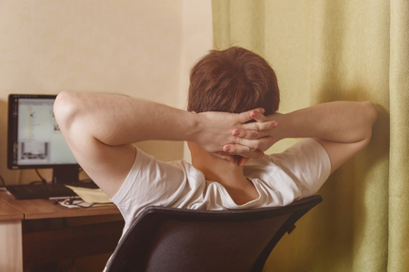 Rear view of man relaxing on chair at home near computer after hardworkの写真素材