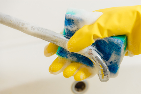 Cleaning Bathroom Faucet with Detergent in Yellow Rubber Gloves with Blue Sponge - Housework, Spring Cleaning Concept.の写真素材