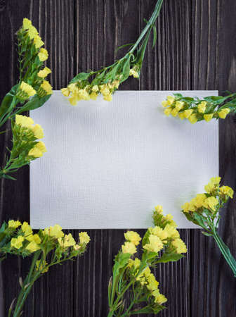 Blank white card decorated with yellow statice flowers on brown wooden background. Mockup. Flat layの写真素材