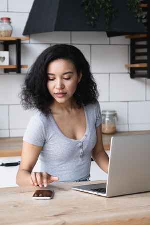 Portrait of attractive African American woman sitting at the kitchen table with a laptop and using a...
