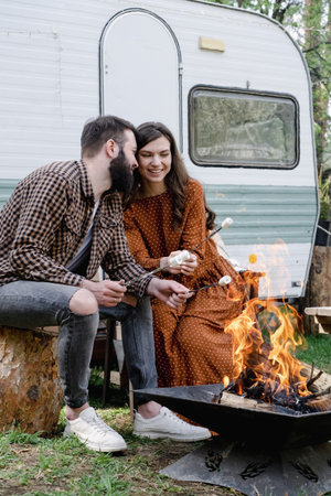 Happy young caucasian couple is traveling in travel van. Romantic atmosphere of relaxation. Road trip around country for weekend. Man and woman roasting marshmallows on campfire. Millennial generationの写真素材