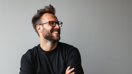 A young man with a beard and glasses is wearing a black t-shirt, smiling and looking away from the camera. The background is a light gray colorの素材