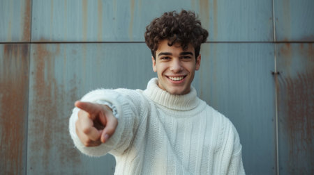 Cheerful young man with curly hair pointing at the camera with a toothy smile on a rusty blue-gray backgroundの素材