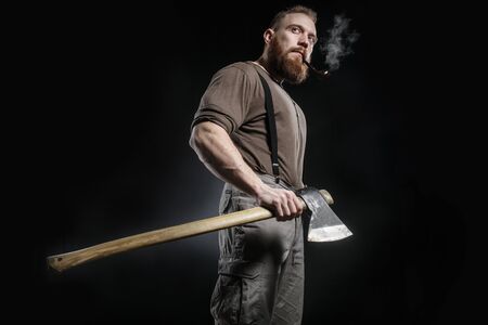 Lumberjack brutal bearded muscled man in brown shirt with smoking tube and suspenders on grey pants...