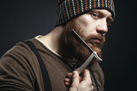 Lumberjack brutal bearded muscled man in brown shirt with shaving blade near his face  on dark backgroundの写真素材