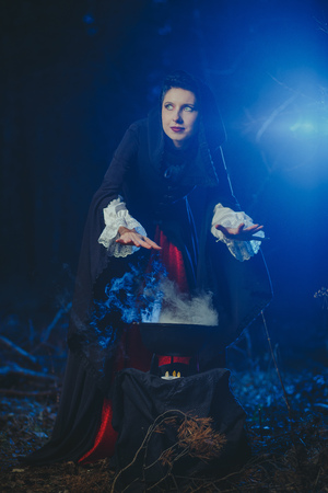 Halloween Young beautiful and mysterious woman witch in woods, in black cloak with hood, witch conjures in the wizarding forestの写真素材