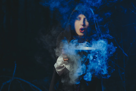 Halloween Young beautiful and mysterious woman witch in woods, in black cloak with hood, witch conjures in the wizarding forestの写真素材