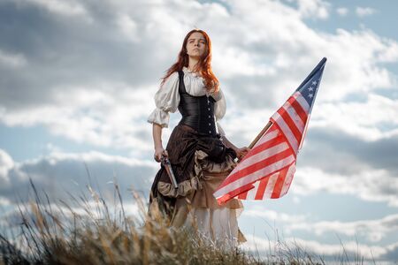 Girl in historical dress of 18th century with flag of United States. July 4 is US Independence Day. Woman of patriot freedom fighter in outdoor on background cloudy skyの写真素材
