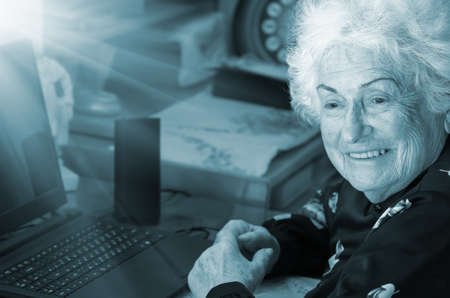 Happy grandmother sits at the table with a computer before learning to use the Internet, blue tone