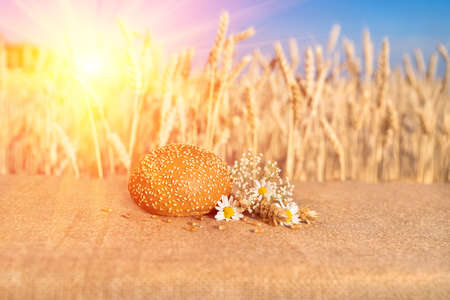 bread bun with sesame seeds lying on a table with flowers of daisies and ears of wheat against the...