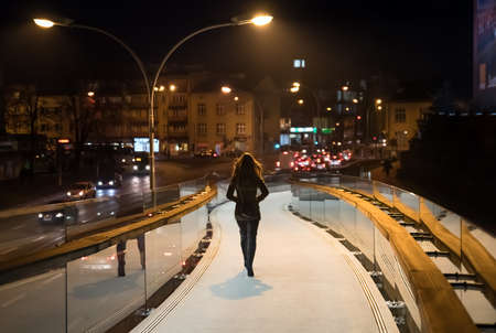 Back view of young brunette woman, going late night on the pedestrian mirror bridge in city Rzeszow in city centre, country Poland, soft focus.の写真素材