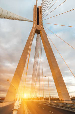 Highway going through a cable-stayed bridge with big steel cables, close-up in the evening during a sunset against a background of sky, clouds and sun raysの写真素材