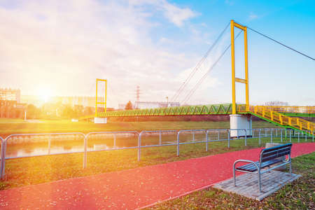 Bench in a park area, near a river and a pedestrian bridge, where people jogging on a rubber...