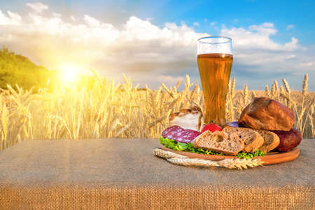 Beer, meat products. Smoked ham, sausage, bacon, cheese, vegetables lying on a table in a wheat field against the blue skyの写真素材