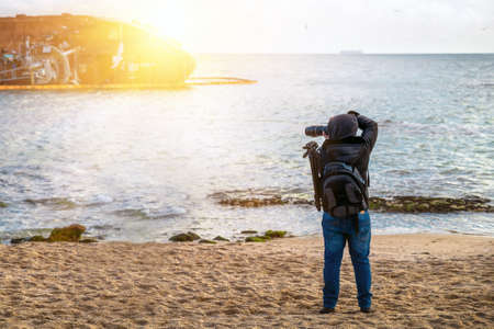 Photographer traveler with a camera and a backpack on the beach in spring when it is still cold, takes pictures sea.の写真素材